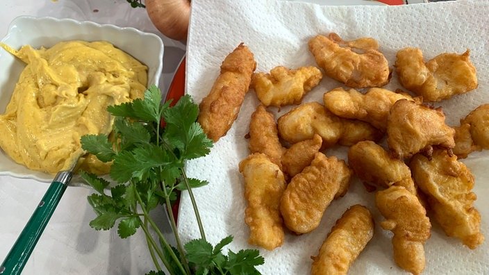 Rechts liegen viele panierte, gebackene Kibbeling auf einem Teller. Links daneben eine Schüssel mit goldgelber Creme.