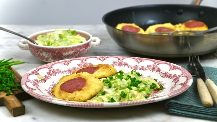 Im vorderen Teil des Bildes ein Teller mit zwei kleinen goldenen Kartoffeltalern und Salat. Im hinteren Teil eine Schüssel und eine Bratpfanne.