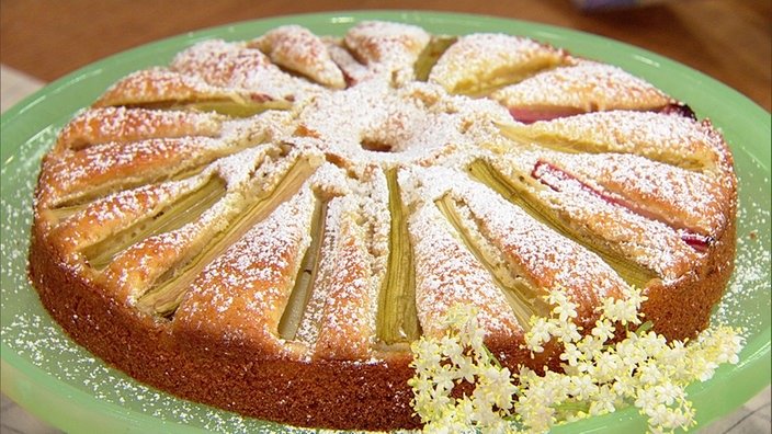 Ein runder Rührkuchen, auf dem aus Rhabarber-Streifen eine Blüte gelegt wurde. Mit Puderzucker berieselt.