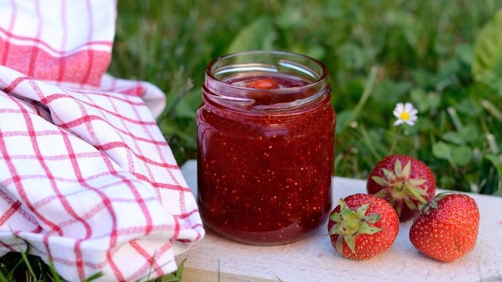 Auf einer Wiese steht ein Glas Erdbeermarmelade. Daneben 3 Erdbeeren zur Dekoration.