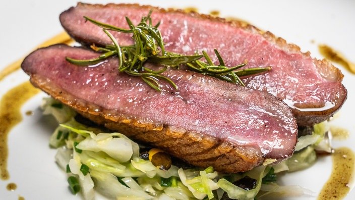 Ein Stück Entenfleisch halbiert und mit Rosmarin garniert. Es glänzt rosa.