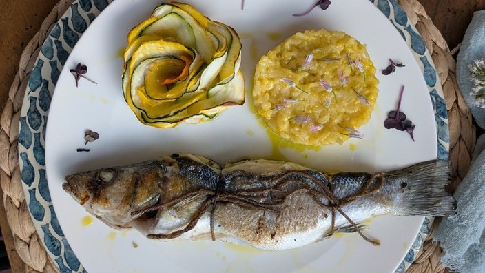 Topshot. Auf einem weißen Teller liegt ein ganzer Fisch. Daneben eine Rose aus Zucchini und ein Klecks Risotto.