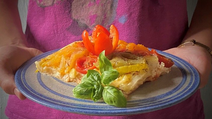 Eine Person hält einen Teller in die Kamera. Darauf ein Stück gelbgoldener Kuchen mit Tomaten und Basilikum garniert. 