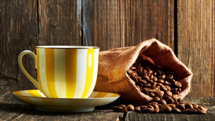Auf einem offenen Kaffeesack purzeln die Kaffeebohnen. Daneben eine gelbweiß-gestreifte große Kaffeetasse mit Unterteller.