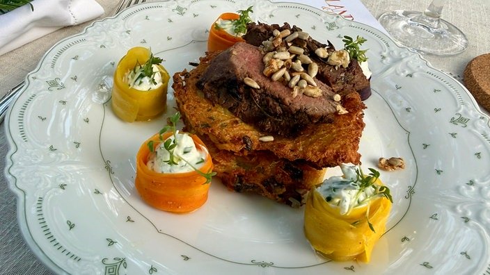 Hübsch drappiert: Auf einem Reibenkuchen liegt ein Stück Fleisch. 3 runde Karottenröllchen mit Frischkäse gefüllt liegen drumherum.