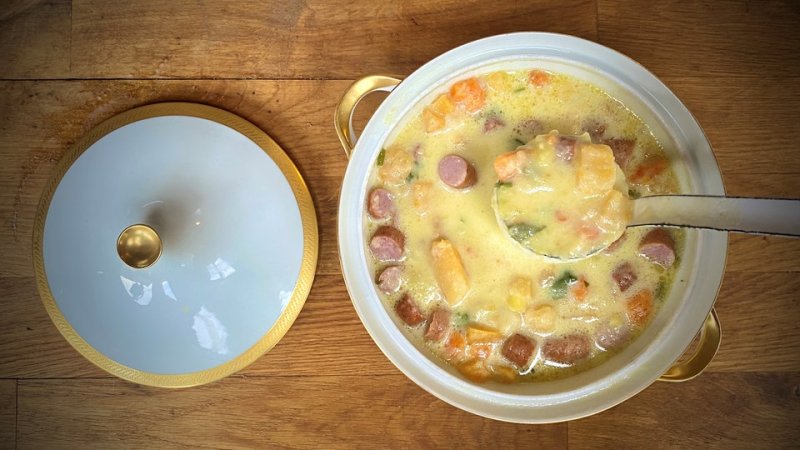 Ein klassischer Suppentopf ist bis oben hin gefüllt mit einer hellen Suppe. Darin schwimmen unter anderem Würstchenstücke.