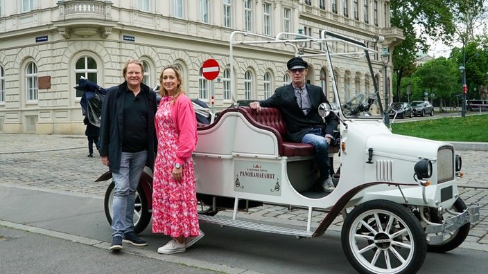 Tamina und Björn stehen neben einer weißen, motorisierten Kutsche vor einem imposanten Gebäude. In der Kutsche sitzt noch ein Chaffeur.