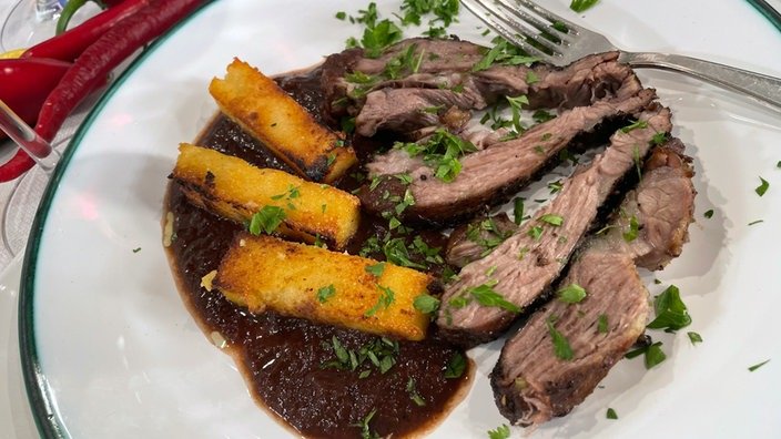 Der Braten liegt in Scheiben auf einem Teller. Garniert mit Petersilie. Daneben Polentaschnitten und ein Klecks braune Soße.