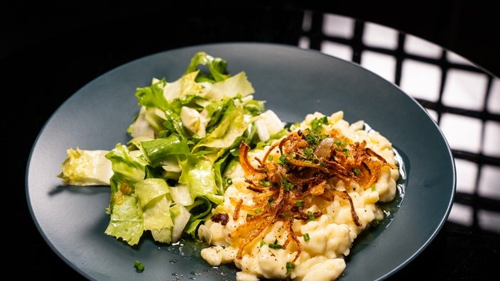 Ein grauer Teller mit einem Klecks Käsespätzle, die mit Zwiebeln garniert sind. Daneben ein Salat.