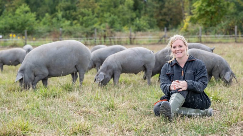 Eine junge Landwirtin auf einem Feld, im Hintergrund mehrere dunkle Iberico-Schweine.