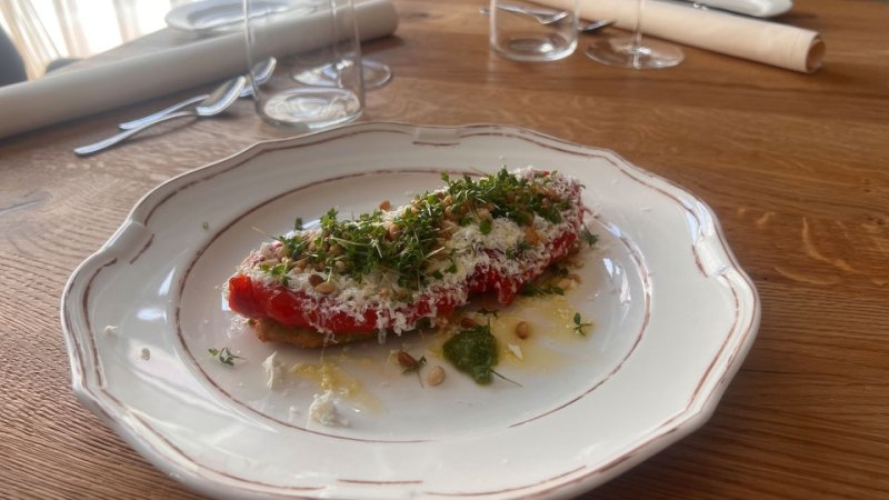 Eine Scheibe Aubergine mit Paprika, Parmesan und Pesto auf einem Teller, garniert mit Kresse und Pinienkernen.