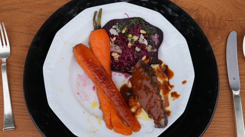 Ein Nackensteak liegt zusammen mit Kartoffelstampf, roter Bete und Möhren auf einem weißen Teller.