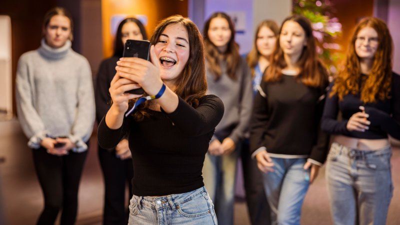 Eine Jugendliche hält ein Smartphone vor sich und filmt oder fotografiert, während mehrere weitere Jugendliche in einem hell beleuchteten Innenraum im Hintergrund stehen.