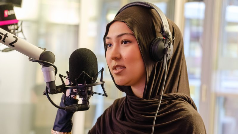 Eine junge Frau mit Kopfhörern spricht in ein Studiomikrofon und steht in einem hellen Radiostudio vor großen Fenstern.