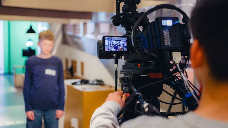 Eine Person filmt mit einer professionellen Videokamera einen Jugendlichen, der in einem Fernsehstudio vor der Kamera steht.