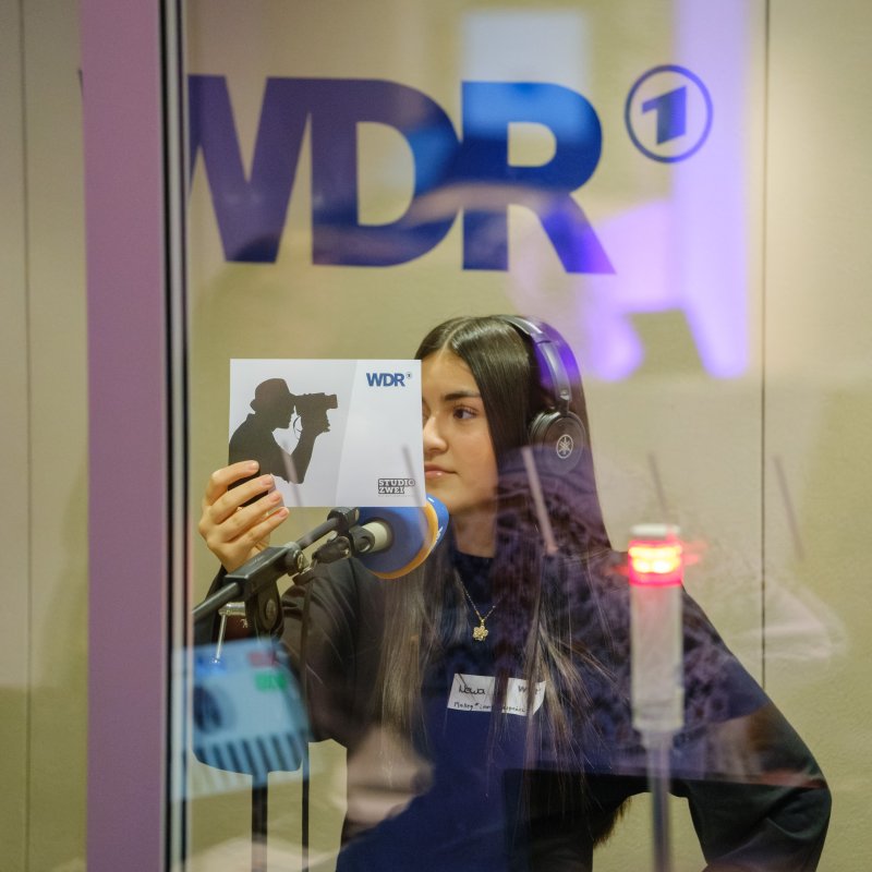 Eine Jugendliche sitzt mit Kopfhörern vor einem Mikrofon im WDR-Studio und hält eine Karte mit Kameramotiv in der Hand; im Hintergrund ist das WDR-Logo zu sehen.