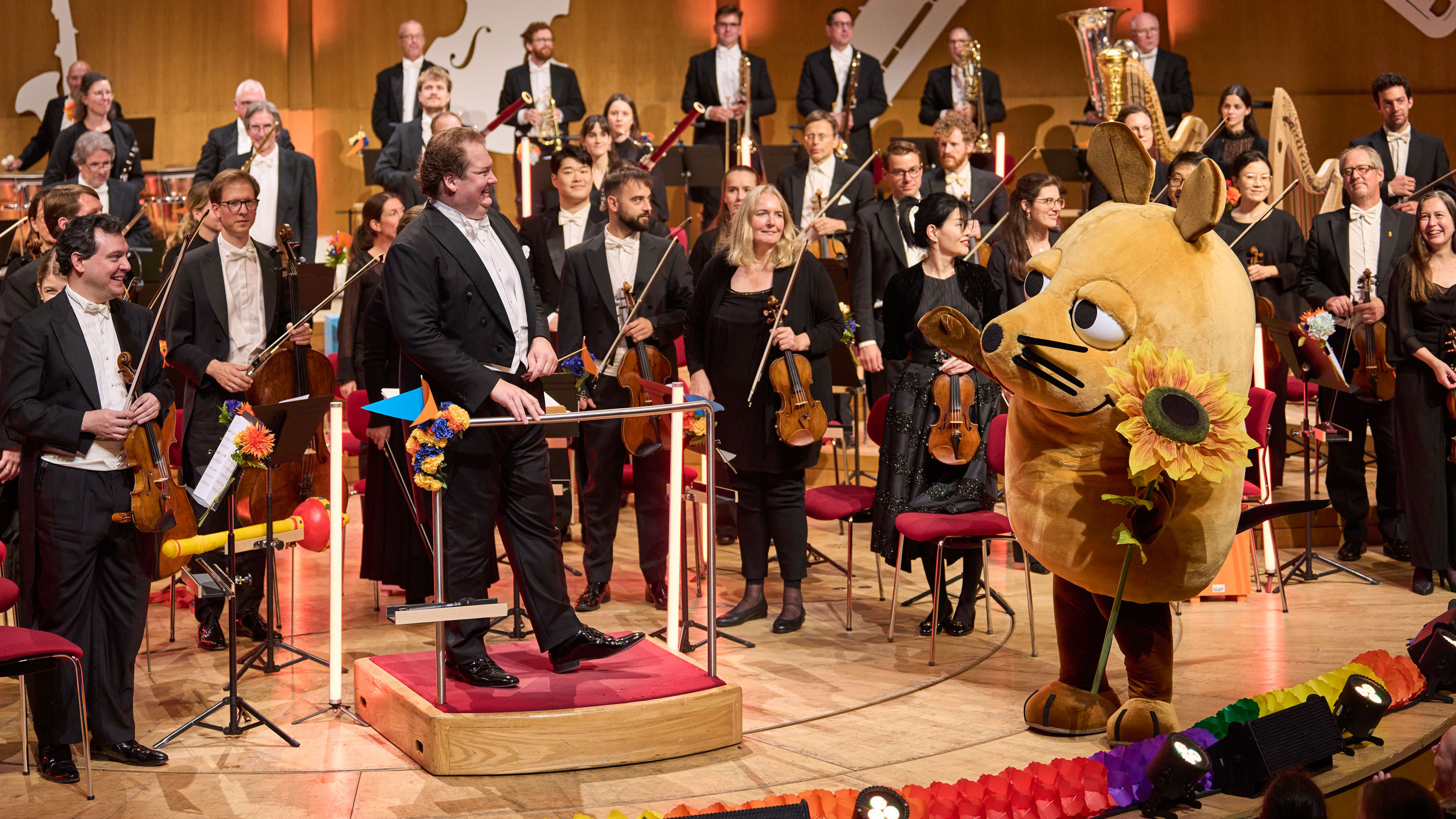 Das Bild zeigt ein klassisches Orchester auf einer Konzertbühne, in dessen Mitte die bekannte „Maus“ aus der „Sendung mit der Maus“ steht und dem Dirigenten eine Sonnenblume überreicht.
