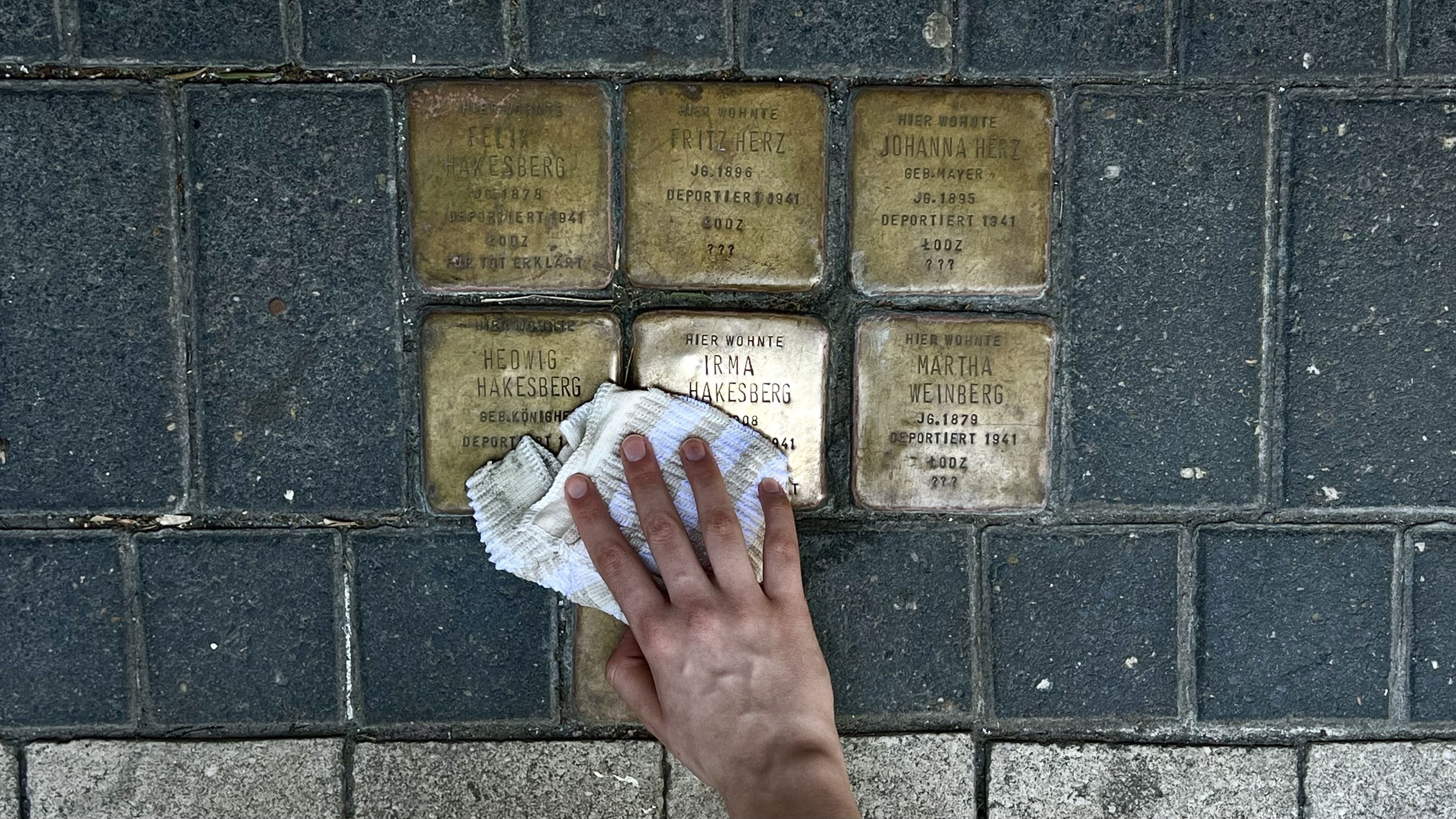 Auf dem Bild ist eine Hand zu sehen, die mit einem Tuch Stolpersteine auf dem Gehweg reinigt.