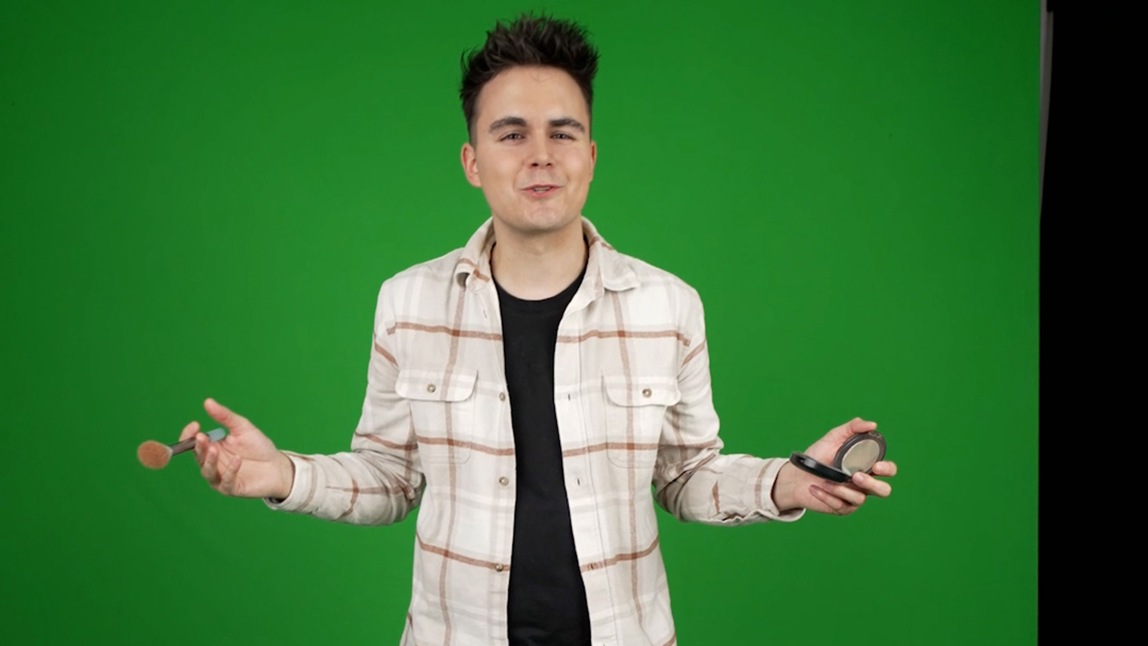 Ein junger Mann steht vor einem Greenscreen, hält einen Pinsel und ein Make-up-Produkt in den Händen und spricht in die Kamera bei einer Videoproduktion.
