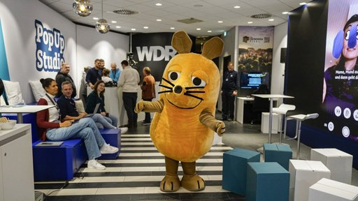 In einem Innenraum, der als "PopUp Studio" und "WDR" gekennzeichnet ist, steht eine Maus in der Mitte des Bildes, während mehrere andere Personen im Hintergrund miteinander interagieren.