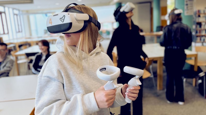 Ein junges Mädchen mit blonden Haaren trägt in einer hellen, schulähnlichen Umgebung ein Virtual-Reality-Headset und hält zwei weiße Controller in den Händen, während weitere Personen im Hintergrund ebenfalls VR-Headsets tragen.