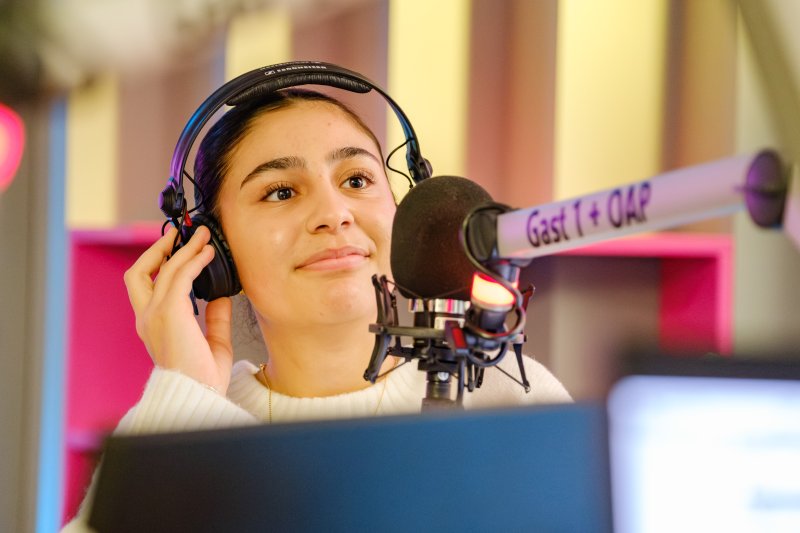 Das Bild zeigt ein Mädchen in einem Studio, das Kopfhörer trägt und vor einem Mikrofon mit der Aufschrift „Gast 1 + OAP“ sitzt.