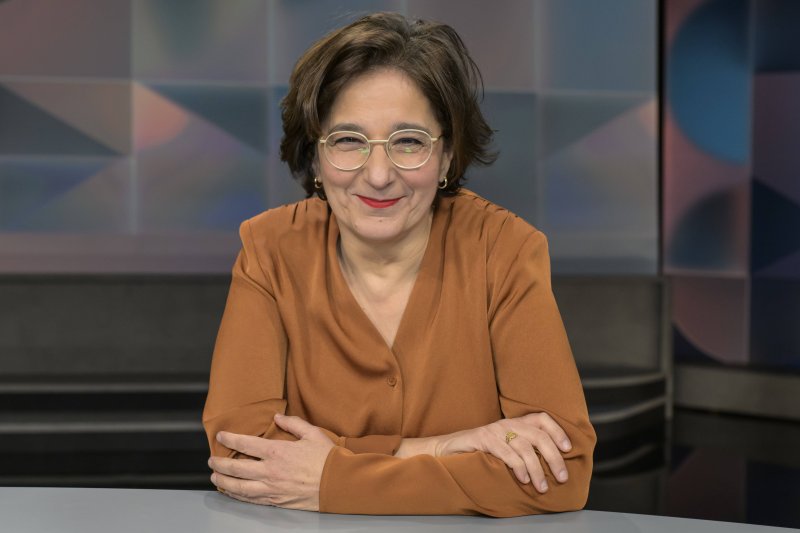 Das Bild zeigt eine Person in einem orangefarbenen Oberteil, die mit verschränkten Armen an einem Studiotisch sitzt.