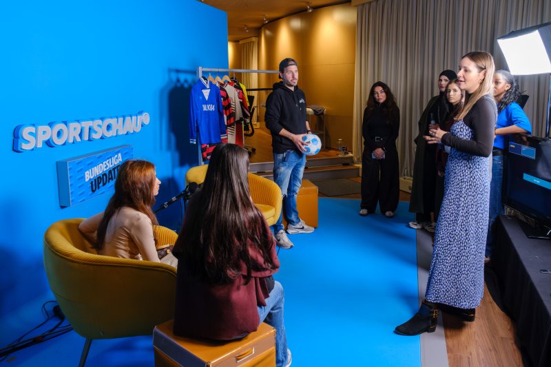Das Bild zeigt eine Gruppe von Menschen in einem blau ausgeleuchteten Raum, in dem ein Sportschau‑Set mit Sitzbereich, Kleidung und Beleuchtung aufgebaut ist.