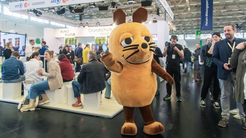 Die orangefarbene Maus läuft als lebensgroßes Maskottchen winkend durch eine große Messehalle, in der viele Menschen an Informationsständen sitzen und stehen.