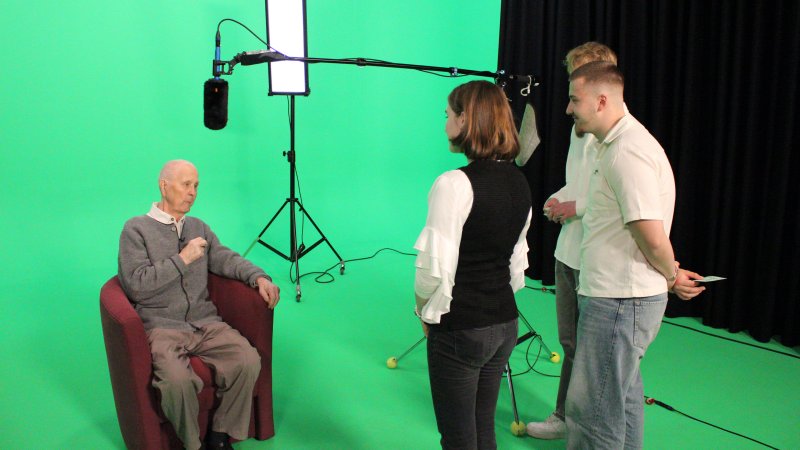Ein älterer Herr sitzt in einem Sessel vor einer Greenscreen-Wand und wird von einem jungen Team für ein Zeitzeugen-Projekt interviewt.