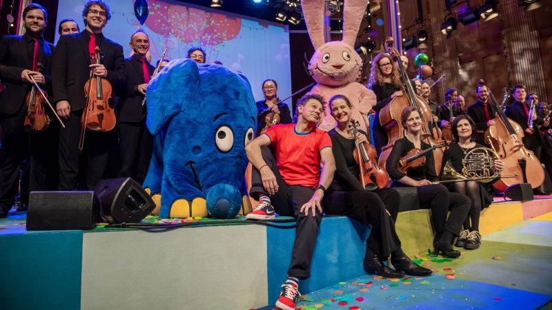Ein Orchester posiert auf einer bunten Bühne zusammen mit Moderator André Gatzke und den bekannten Figuren Elefant und Hase aus der "Sendung mit dem Elefanten".